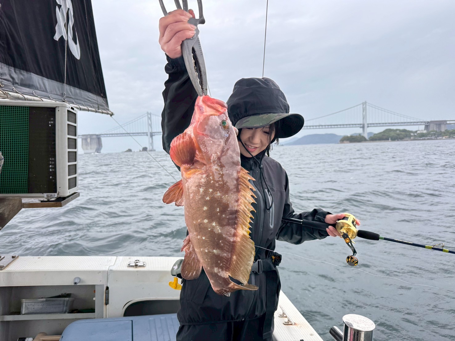 Person on a boat proudly holds up a large reddish-brown fish they just caught, wearing a black rain jacket and hat with a fishing rod in the other hand, water and a bridge in the background.