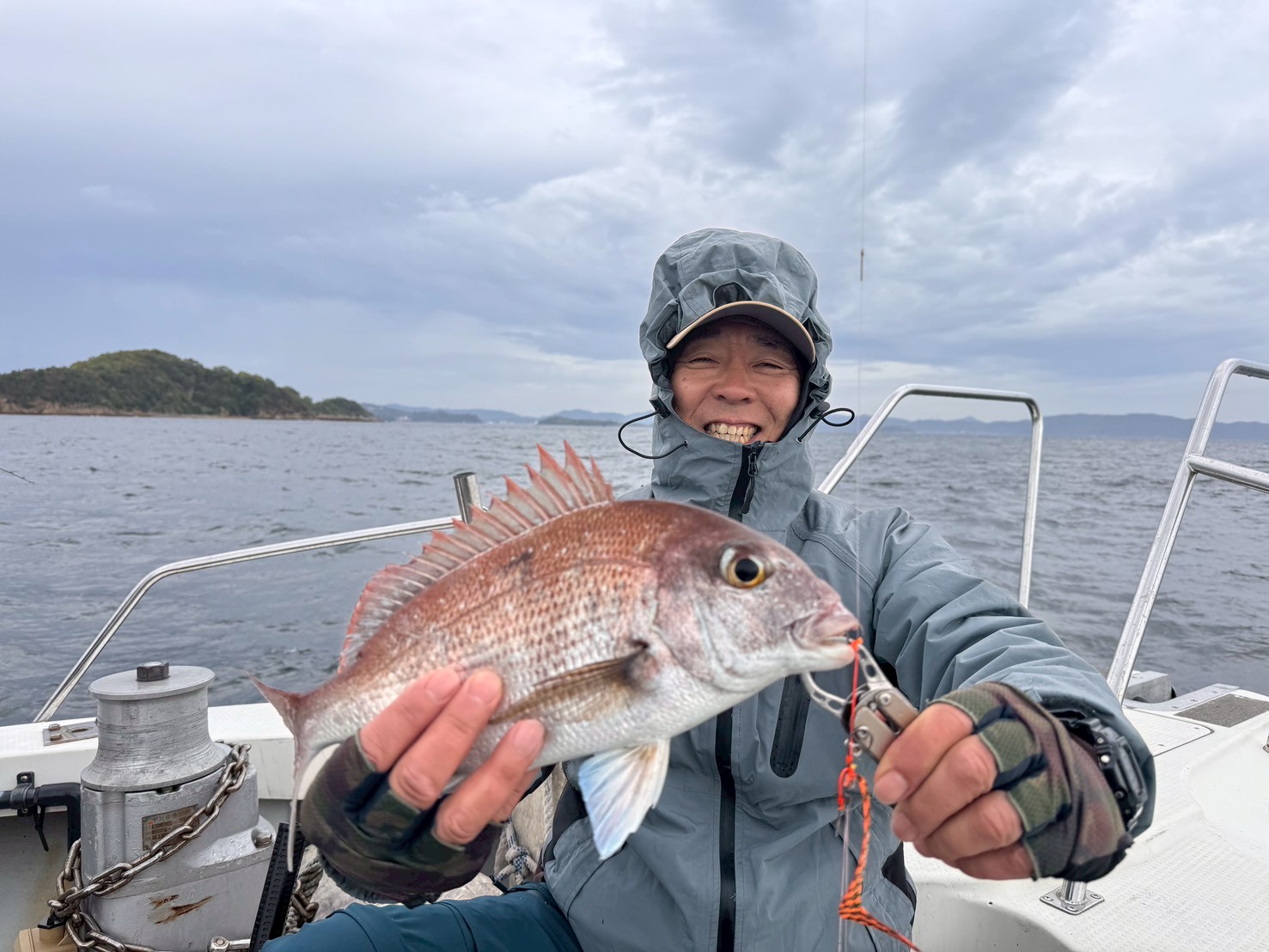 Person in a hooded rain jacket on a boat holding a red snapper with a lure in its mouth.