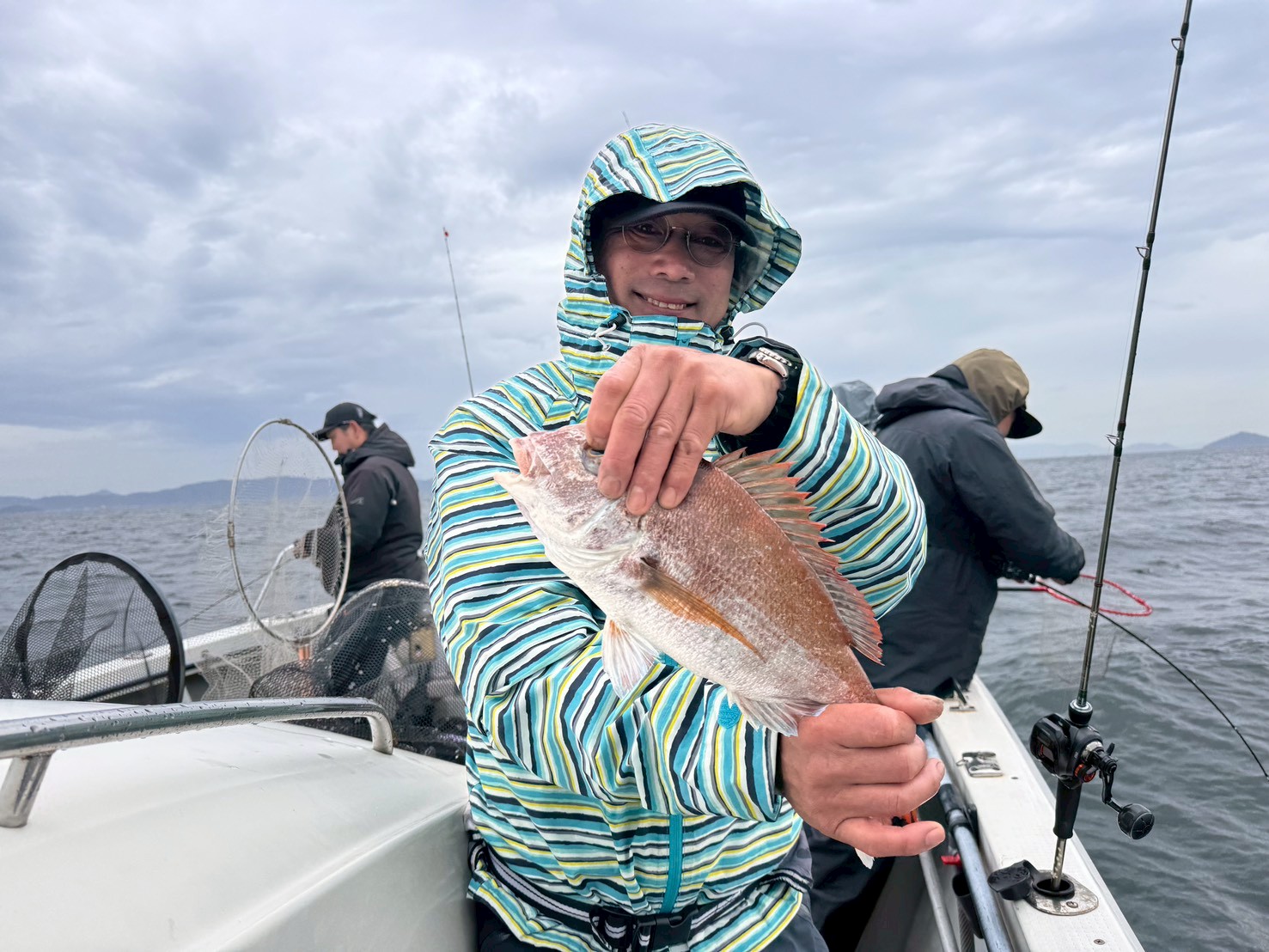 Angler on a boat holding up a pinkish-red fish to the camera, with other fishermen fishing in the background on a choppy sea under a cloudy sky.