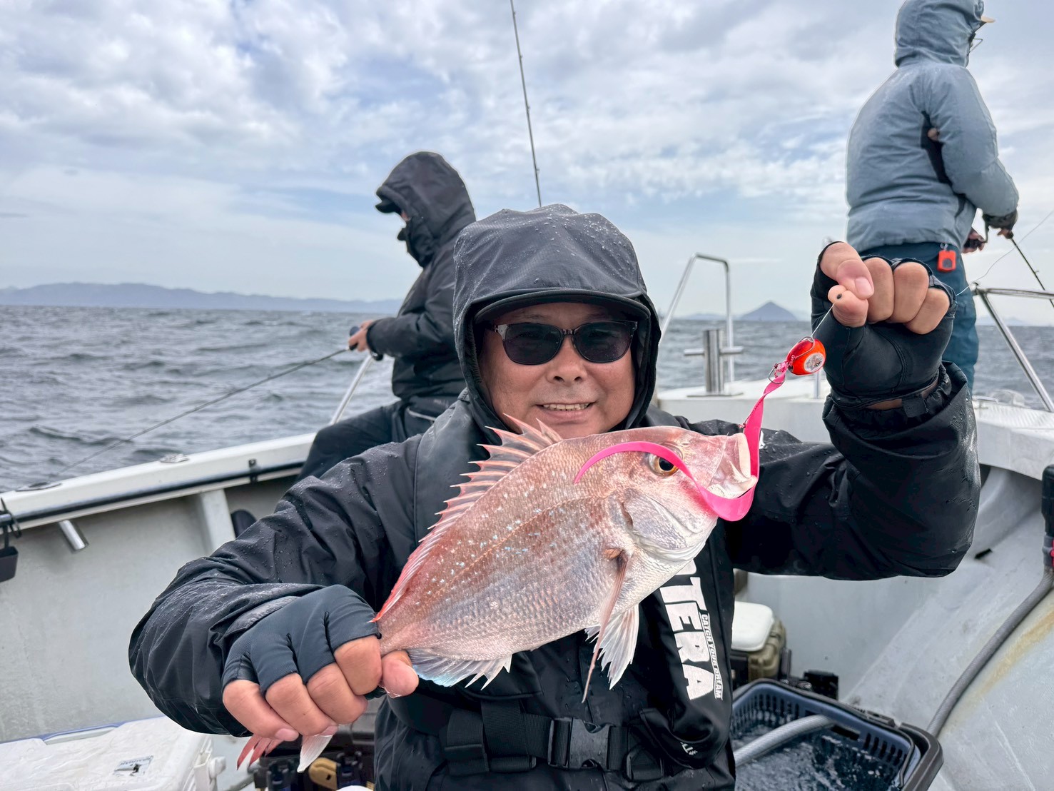 Person in a hooded jacket on a boat holds up a pink fish caught on a bright lure with the ocean in the background.