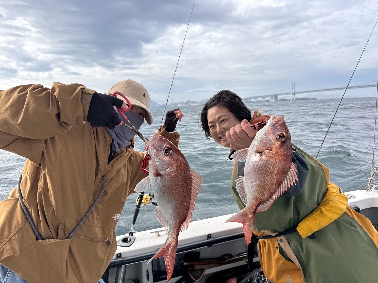 Two people in rain jackets on a boat proudly hold up two red fish they just caught, with the ocean and a distant bridge in view.