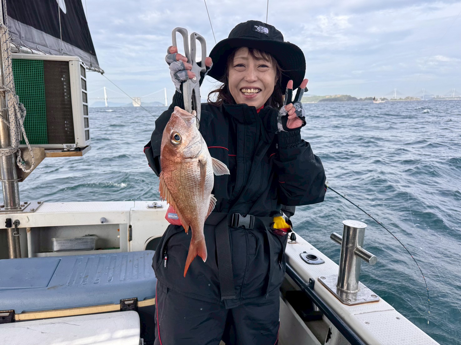 Smiling woman on a fishing boat holds up a red snapper with pliers, ocean behind.