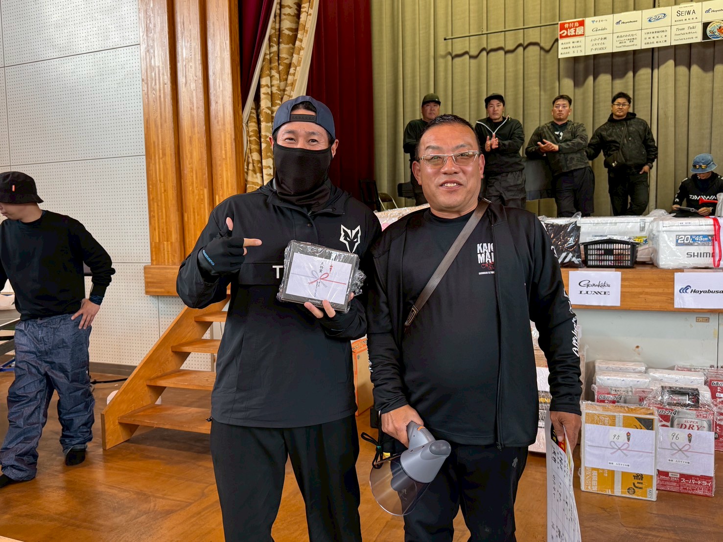 Two men in black outfits pose for a photo at an indoor event; the left man holds a wrapped prize and gives a thumbs up.