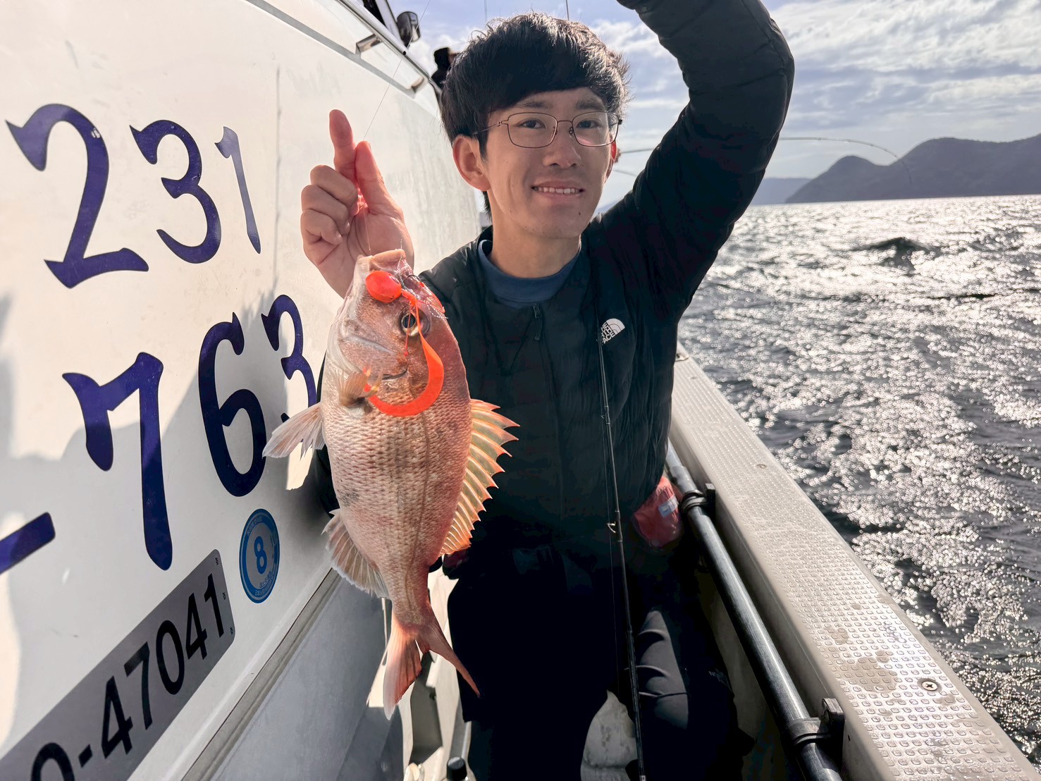 Person on a boat holds up a reddish fish caught with an orange lure, with water and distant hills in the background.