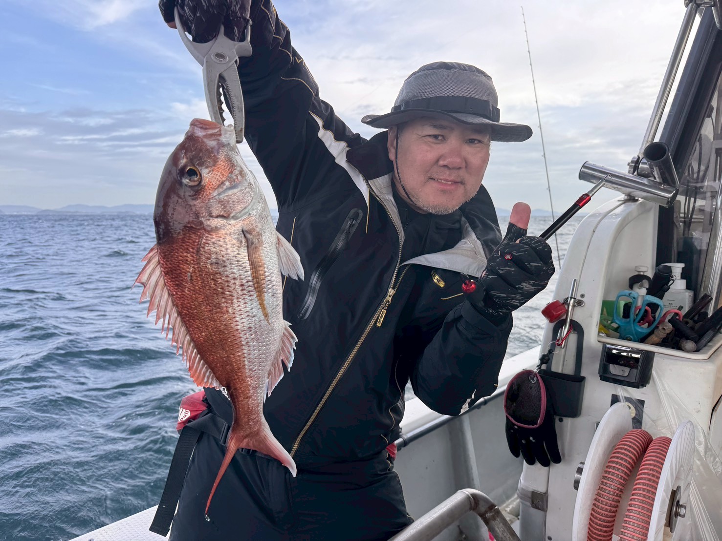 Fisherman on a boat proudly holds a red snapper with a fish gripper, giving a thumbs-up by the rail.