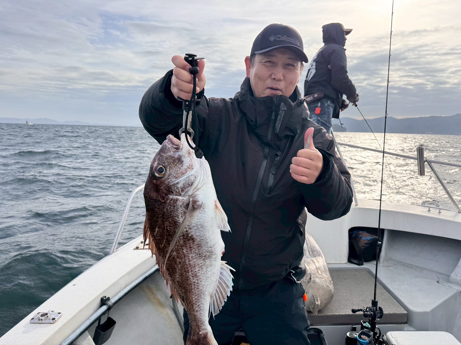 Man in a black jacket on a boat holds a large fish with a fish grip, giving a thumbs up to the camera.