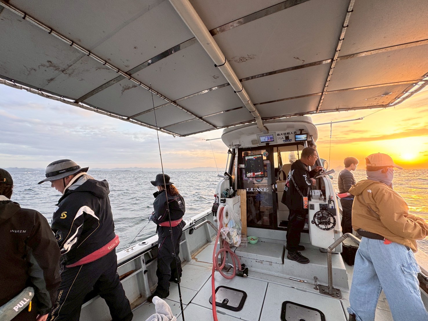 Fishermen standing along the rail and casting lines from a covered boat at sunset over calm water, distant horizon visible.