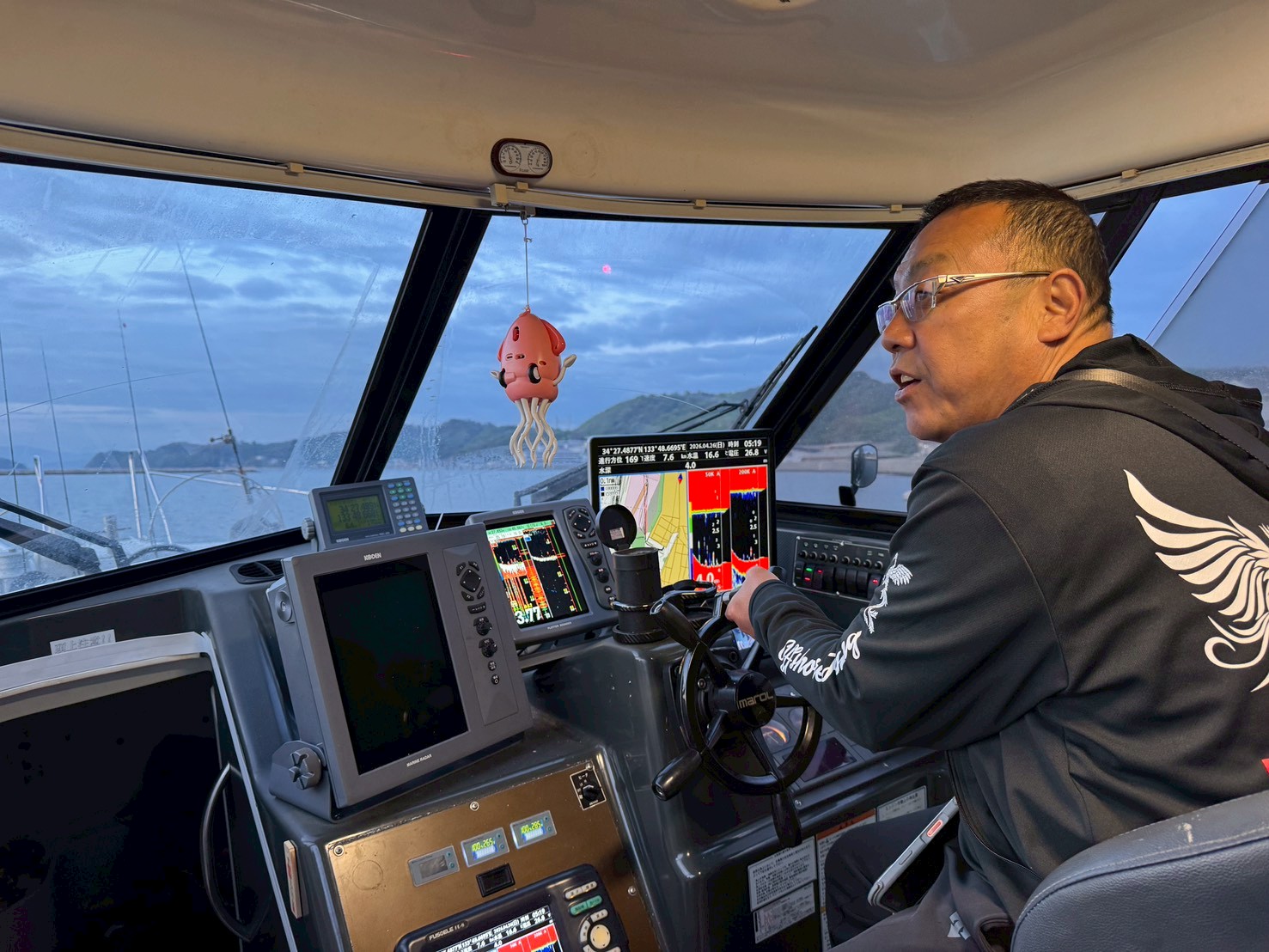 Captain at the helm inside a boat cockpit, monitoring multiple navigation screens as the sea and distant coastline appear through the windows, with a pink octopus ornament hanging above.