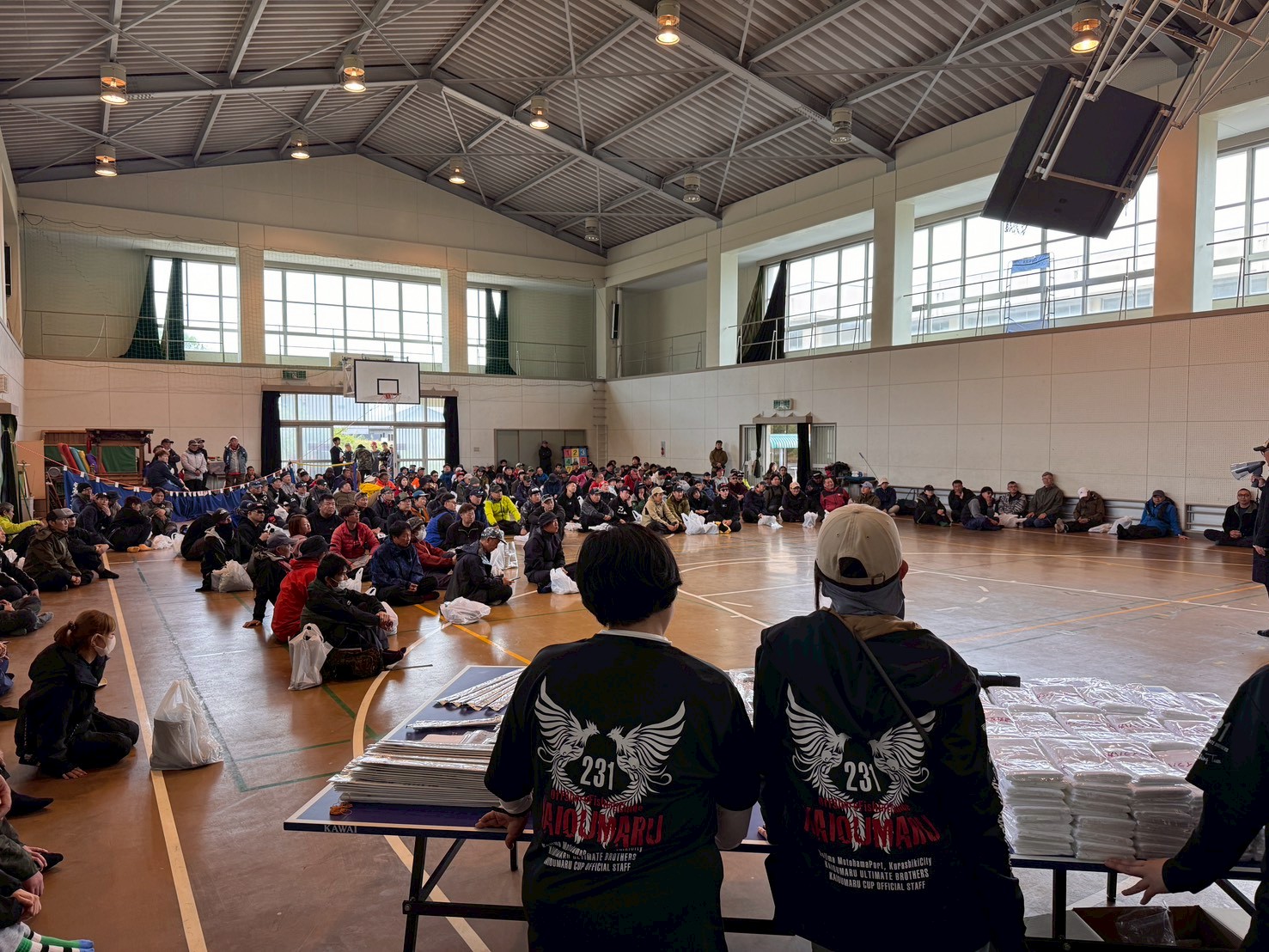 Wide view of a crowded school gym with attendees seated on the floor, facing a stage area where two organizers in black shirts stand behind a table with wrapped items and papers in the foreground.