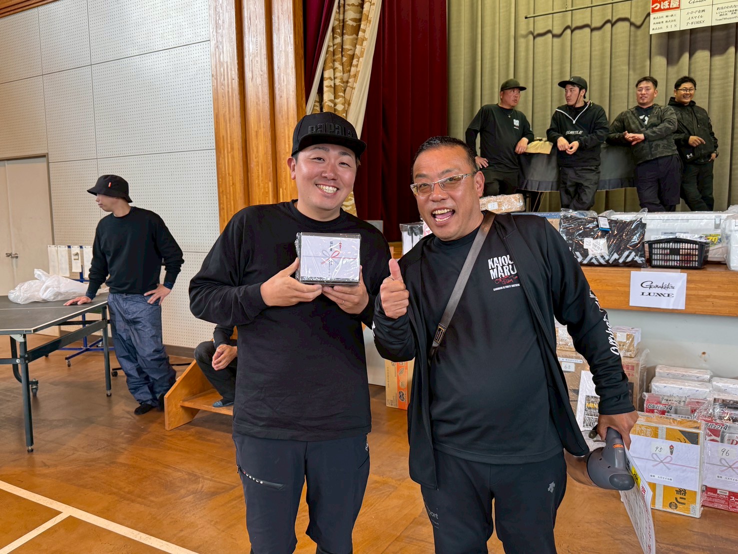 Two men in black stand at the camera; one holds a wrapped prize while the other shows a thumbs‑up in a gym/assembly hall setting.