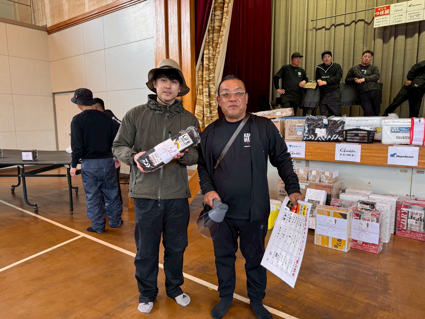 Two men pose for a photo in a gym event; one holds a wrapped prize, the other carries a clipboard and protective gear, with prize boxes on the stage behind them.