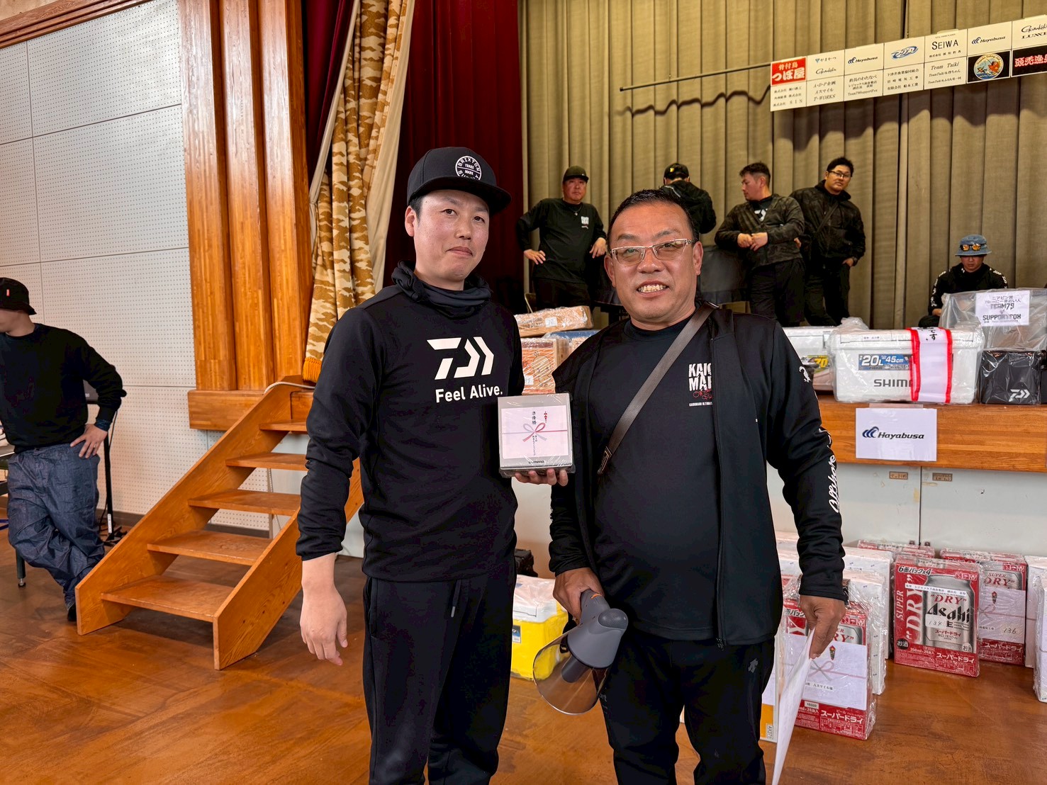 Two men in black athletic wear pose together in a hall, one holding a wrapped gift while others and prize boxes sit on a stage behind them.