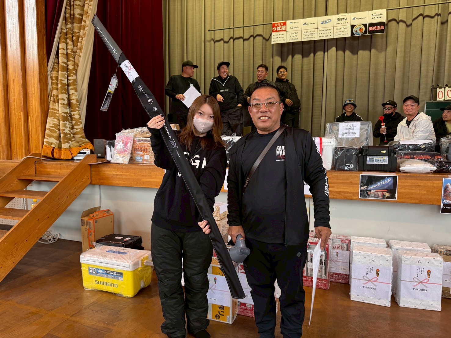 Two people stand in a hall on a stage area; the woman in a mask holds a long black item like a fishing rod case beside a man in black smiling at the camera.