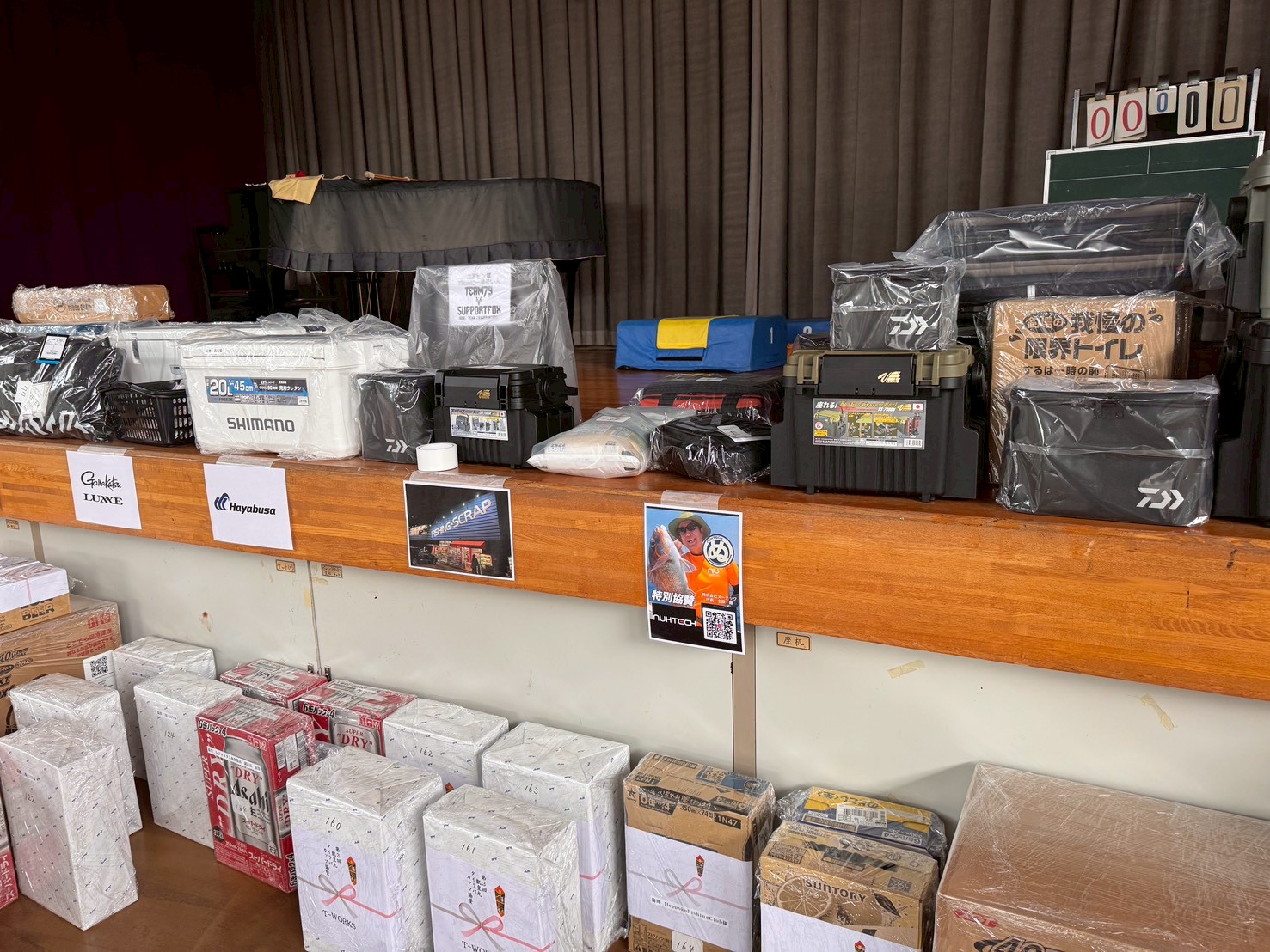 Row of wrapped fishing gear and storage boxes on a stage, with brands like Shimano and Hayabusa visible on the containers